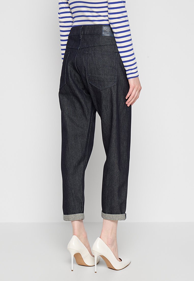 Femme portant un jean en denim noir retroussé et des talons hauts blancs, avec un haut à rayures bleu et blanc à manches longues, se tenant devant un fond uni.