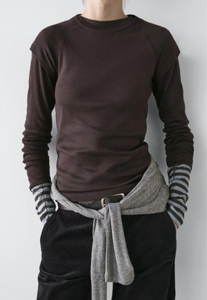 Personne portant une chemise à manches longues marron foncé par-dessus des manches rayées grises et noires, un pull gris noué à la taille, et un pantalon noir avec les mains dans les poches.