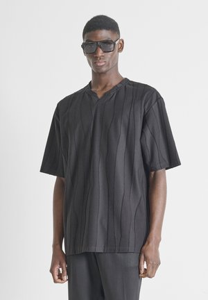 Joven con camisa negra oversized con sutiles rayas verticales y grandes gafas de sol negras, de pie frente a un fondo blanco.