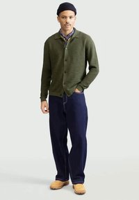 Muž v tmavě zeleném zapínacím cardigan, modrých širokých džínách, hnědých botách, černé čepici a vzorovaném šátku kolem krku, stojící s jednou rukou v kapse.