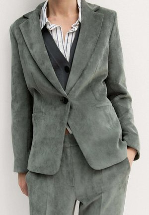 Persona che indossa un completo di velluto a coste verde con blazer e pantaloni abbinati sopra una camicia a righe e un gilet scuro, mano in tasca.