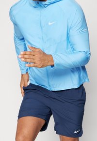 Coupe-vent bleu clair Nike avec capuche, fermeture éclair et détails réfléchissants, associé à un short bleu marine avec un logo blanc. Position de course.