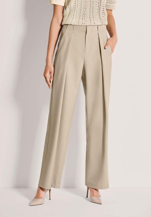 Beige Hosen mit weitem Bein und vorderen Falten, mit zwei seitlichen Taschen, maßgeschneidertem Design und knitterfreiem Stoff, kombiniert mit hellfarbigen spitzen Schuhen.