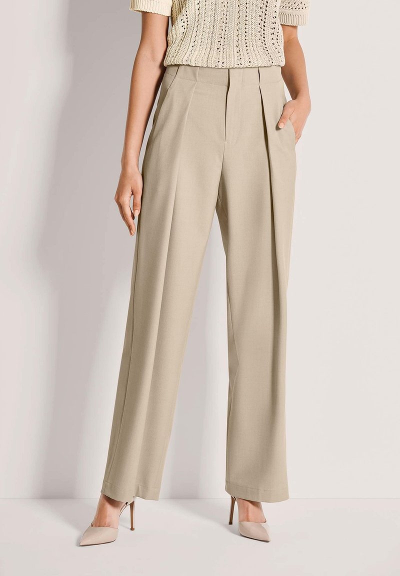 Beige Hosen mit weitem Bein und vorderen Falten, mit zwei seitlichen Taschen, maßgeschneidertem Design und knitterfreiem Stoff, kombiniert mit hellfarbigen spitzen Schuhen.