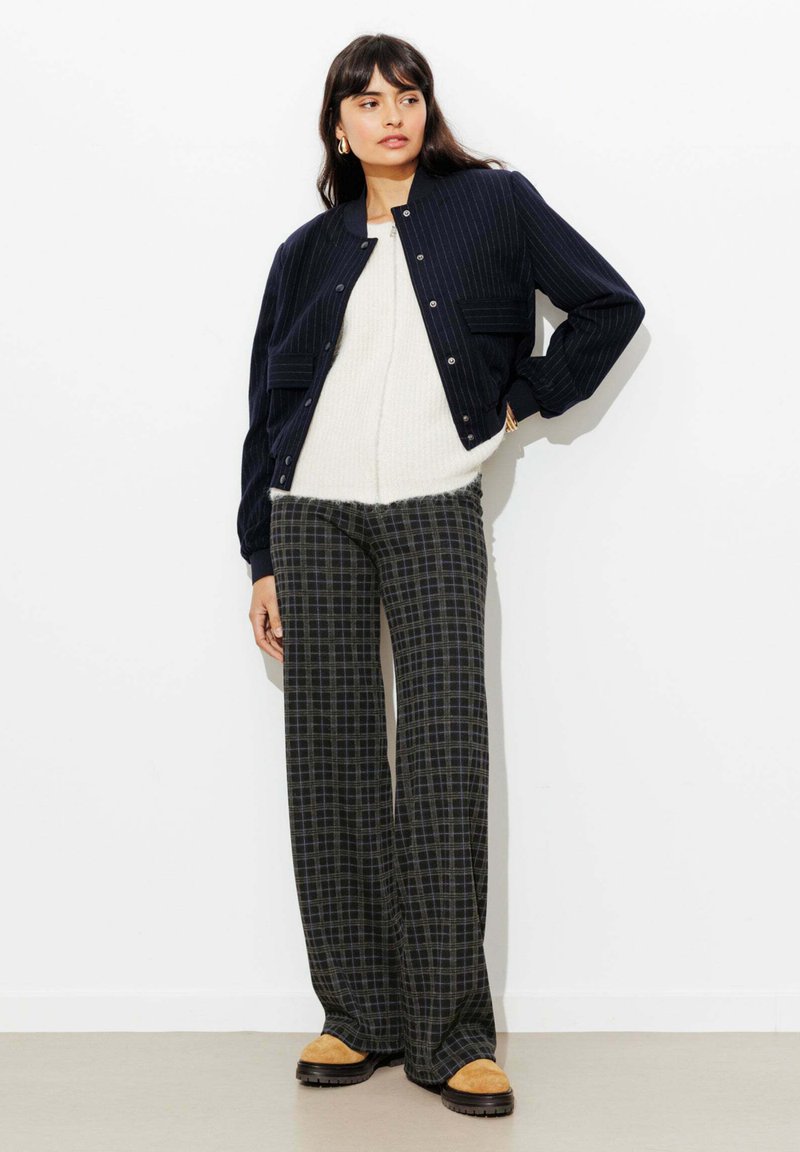 Veste aviateur à fines rayures bleu marine, pull crème en tissu duveteux, pantalon large à carreaux gris et chaussures à enfiler couleur beige. Design minimaliste avec une coupe structurée.