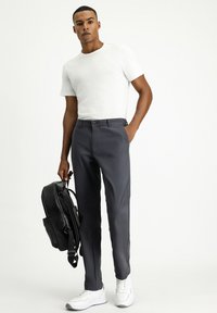 Pantalones gris tapered con cinturón, combinados con una camisa blanca de manga corta. Una mochila negra se sostiene en una mano. Zapatillas completan el conjunto.