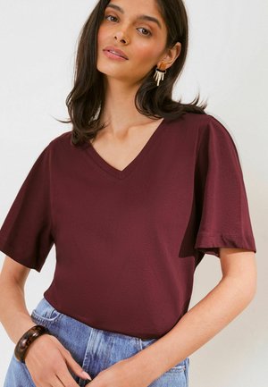 Kvinde iført en vinrød V-hals kortærmet skjorte, blå jeans, et skildpaddeskal-armbånd og geometriske øreringe, poserer med hænderne ved taljen.