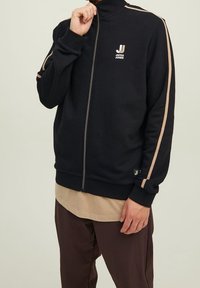 Giacca nera con zip realizzata in materiale morbido, con strisce crema lungo le maniche e un piccolo logo sul petto. Vestibilità rilassata con polsini a coste.