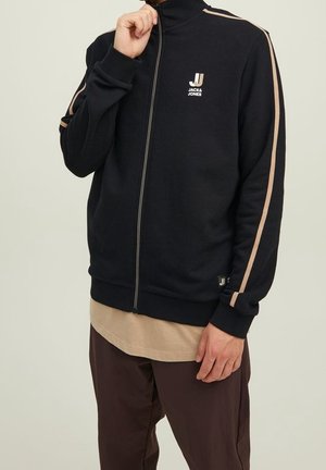 Chaqueta negra con cremallera, hecha de material suave, que presenta rayas crema a lo largo de las mangas y un pequeño logo en el pecho. Corte relajado con puños acanalados.