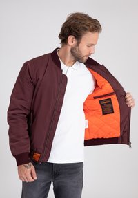 Giubbotto bomber bordeaux con polsini e colletto a coste; fodera arancione trapuntata; tasca interna sinistra con etichetta; indossato sopra una maglietta polo bianca.