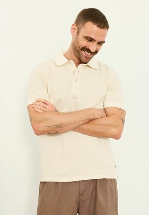 Smiling man with short hair and mustache, wearing a cream polo shirt and brown pants, arms crossed, looking down against a plain background.