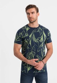 Marineblauwe t-shirt met korte mouwen, voorzien van een groene tropische bladerenprint, met een ronde hals en gemaakt van katoen. Gecombineerd met marineblauwe jeans.