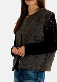 Gilet en laine grise au design carré, avec des manches noires agrémentées de sequins. Tissu texturé et style ouvert à l'avant. Assorti à un pantalon en jean bleu.