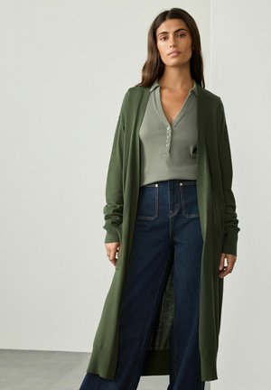 Cardigan lung verde cu deschidere în față, cu manșete ribișate, purtat peste un top henley verde deschis și blugi evazați din denim închis. Material texturat.