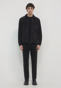 Giovane in piedi, che indossa una giacca nera con zip sopra una maglietta nera, pantaloni neri e scarpe nere, contro uno sfondo chiaro e neutro.