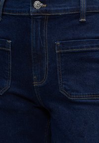 Μπλε σκούρο τζιν μπροστά με ορατό ασημί κουμπί και διπλά ραμμένες τσέπες και ραφές.