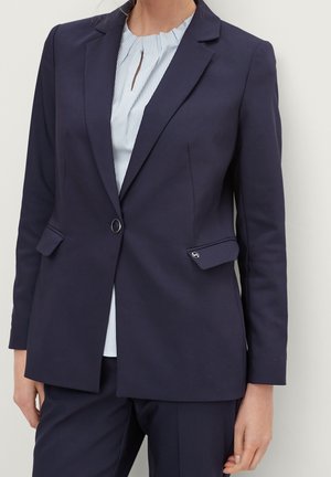 Femme portant un blazer bleu marine foncé avec un seul bouton et des poches à rabat sur un chemisier rayé bleu clair et un pantalon assorti.