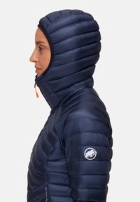 Kvinna i marinblå vadderad jacka med huva och mammut-logotyp på ärmen, vänd mot vänster mot en ljus, enkel bakgrund.