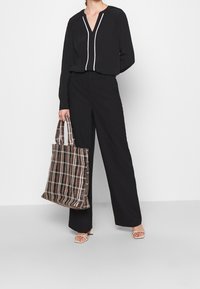 Blusa nera a maniche lunghe con piping bianco, abbinata a pantaloni neri a gamba larga. Borsa a tracolla a quadri marrone tenuta in mano, con doppi manici.