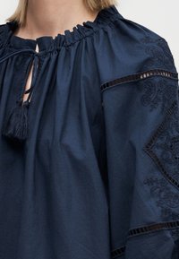 Μπλούζα ναυτικού μπλε με σούρες στο ντεκολτέ, κορδόνια με φούντες και κεντητά λουλουδένια σχέδια στις φουσκωτές μανσέτες με διακοσμητικές λεπτομέρειες από κορδέλα.