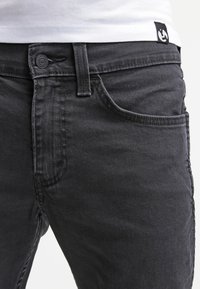 Mustat farkut, joissa on klassinen viiden taskun muotoilu, nappisuljenta ja ommeltu yksityiskohtaisuus. Materiaali näyttää teksturoidulta ja kestävältä.