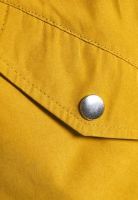 Žlutá látka s hladkým povrchem, obsahující klopovou kapsu zajištěnou stříbrným patentem a detailním prošíváním podél okrajů.