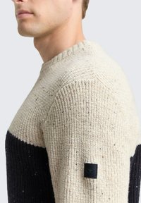 Strickpullover mit einem beigen und marineblauen Farbblockdesign, strukturiertem Stoff mit Farbspritzern und einem schwarzen Logotag am Ärmel.