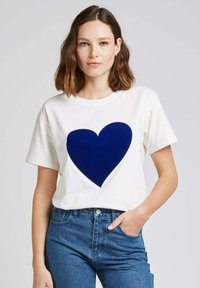 T-shirt en coton blanc avec un grand motif de cœur bleu marine sur le devant. Manches courtes et col rond. Coupe décontractée.