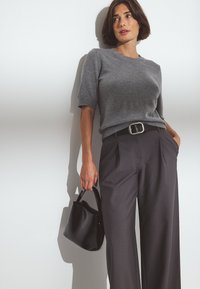 Pelēka adīta īsām piedurknēm blūze, augstās jostas pelēki platās bikses ar melnu jostu, tur rokās maza melna somiņa. Vienkāršs faktūras fons.