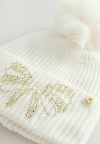 White knitted beanie featuring a fluffy pom-pom, adorned with gold rhinestone bow design and a small gold heart accent.
