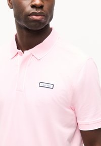 Homme portant un polo Calvin Klein rose clair avec col et manches courtes, cadré aux épaules et au cou.
