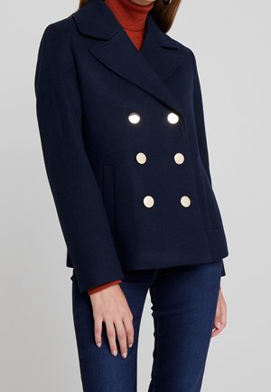 Frau trägt einen marineblauen zweireihigen Mantel mit großen goldenen Knöpfen über einem rostfarbenen Rollkragenpullover und dunkelblauen Jeans.