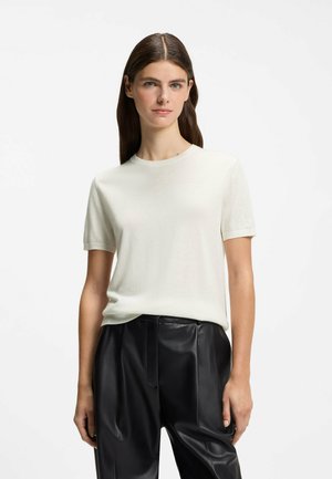 Jeune femme aux longs cheveux bruns portant un t-shirt blanc simple à manches courtes et un pantalon en cuir noir sur fond blanc.