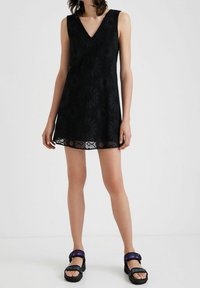 Vestido negro sin mangas con escote en V, que presenta un patrón de encaje floral y un dobladillo con forma de caracol. Combinado con sandalias negras de suela gruesa.