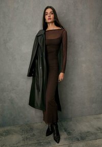 Langes braunes Kleid mit tailliertem Oberteil und transparenten, langen Ärmeln, kombiniert mit einem schwarzen Leder-Trenchcoat. Texturierte, hochhackige Schlangenleder-Stiefel.