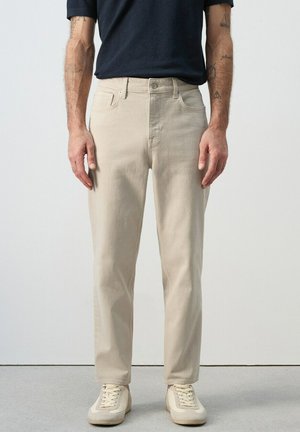 Mann trägt beige gerade geschnittene Hose, cremefarbene Turnschuhe und ein dunkles kurzärmeliges Hemd, steht vor einem schlichten Hintergrund.