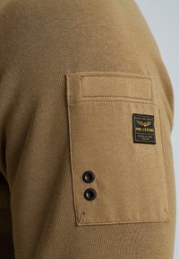 Khaki-Pullover aus strukturiertem Stoff, ausgestattet mit einer aufgesetzten Tasche mit einem schwarzen Etikett und zwei schwarzen Knopfakzenten an den oberen Ärmeln.