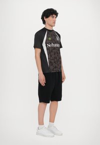 Camiseta atlética preta com sobreposição padronizada, detalhes brancos e texto. Combinada com calções pretos e ténis brancos. Apresenta um design desportivo.