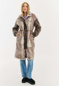 Žena oblečená v dlouhém vzorovaném plášti z umělé kožešiny s opaskem, světle fialovém topu, modrých džínách a černých platformových botách, stojící se zahnutýma rukama v kapsách.