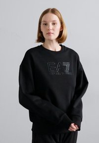 Μαύρη oversized φουτεράκι με στρογγυλή λαιμόκοψη, εμφανίζοντας το ανάγλυφο λογότυπο "EA7 Emporio Armani" σε αντίθεση υφής. Μαλακό ύφασμα, χαλαρή εφαρμογή.