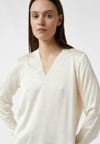 V-Ausschnitt, langärmliges Oberteil aus weichem cremefarbenem Stoff. Verfügt über eine glatte Textur und dezenten Glanz, mit einem klaren, schlichten Design.