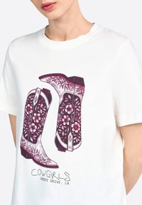 La camiseta blanca presenta un gráfico de dos botas de vaquera con diseños florales en púrpura y rosa, el texto debajo dice "COWGIRLS RODEO DRIVE, LA."