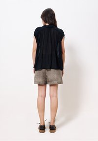 Femme de dos portant un haut noir transparent à manches courtes, un short à carreaux beige et noir, et des chaussures marron à lacets sur un fond blanc uni.