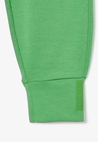 Gröna bomullsjoggers med ribbad textur vid mudden och ett rektangulärt gummimärke med geometriskt mönster. Sömmar i detalj nära mudden.