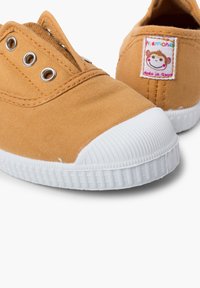 Baskets en toile couleur moutarde avec un embout et une semelle en caoutchouc blanc. Présente des œillets en métal et un logo de singe brodé sur le talon.