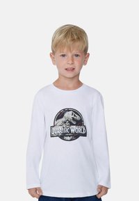 Biała T-shirt z długim rękawem, z graficznym nadrukiem dinozaura i napisem "Jurassic World" w czerni i czerwieni na przodzie.