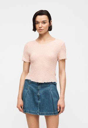 Ung kvinde med kort mørkt hår iført en tekstureret lysepink kortærmet top og en blå denimnederdel, stående mod en ensfarvet baggrund.