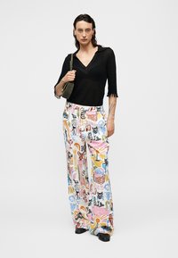 Femme portant un haut noir transparent et un pantalon ample à jambes larges avec des imprimés abstraits colorés et animaliers, tenant un petit sac à bandoulière olive.