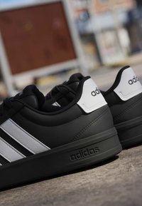 Paire de baskets noires Adidas avec des bandes blanches et un logo, placées à l'extérieur sur une surface en béton.