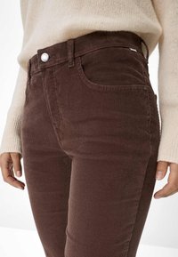Donna che indossa pantaloni in corduroy marroni e un maglione lavorato a maglia color crema, in piedi con le mani rilassate lungo i fianchi.